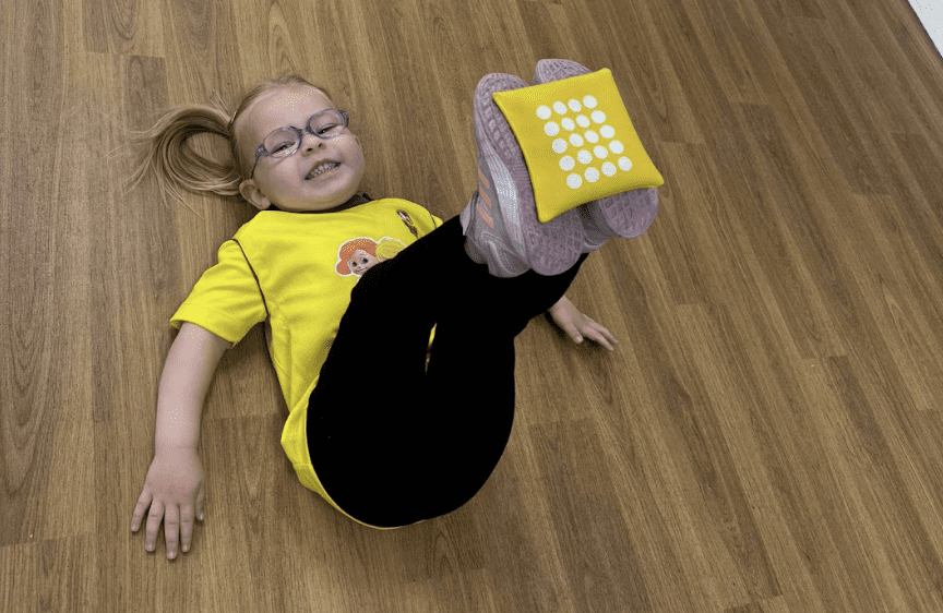 Football confidence for toddlers. Image shows girl laying down balancing a bean bag on her feet in a Socatots class.