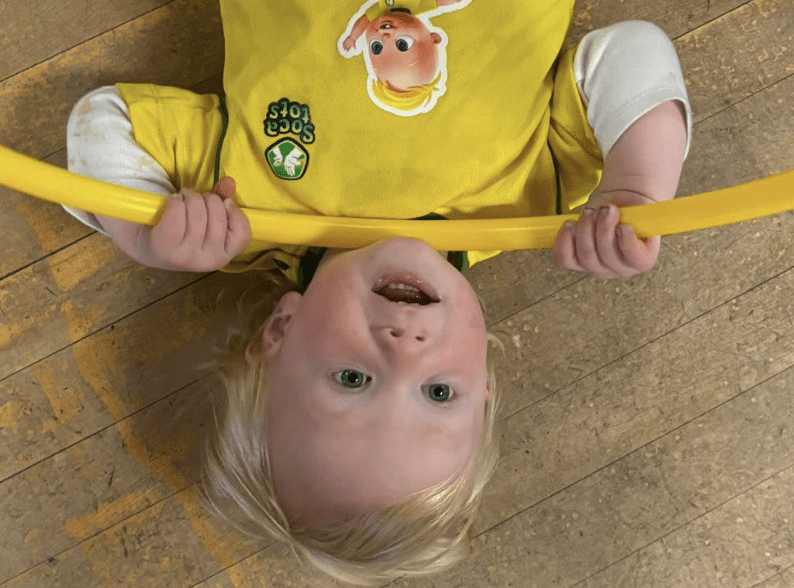 Image shows a toddler on their back with a hula hoop in a Socatots shirt.