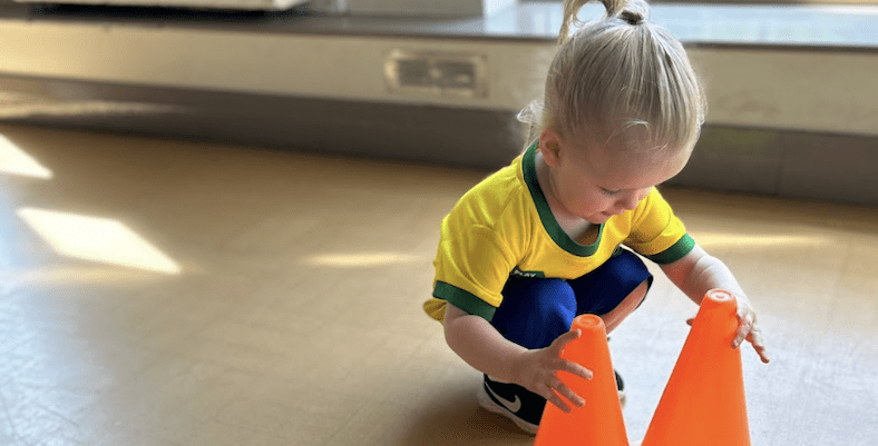 When is a baby a toddler. Image shows a toddler bending down and playing with two orange plastic cones in a socatots class.