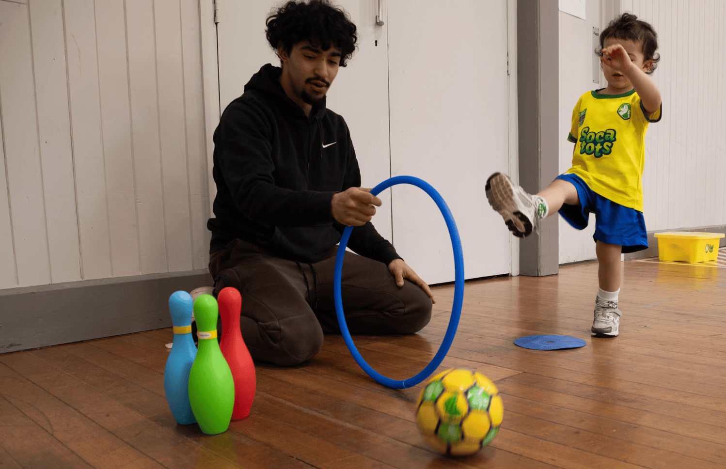 Toddler learning style football. Image shows a father and son at a Socatots class with the son kicking a football through a hoop.