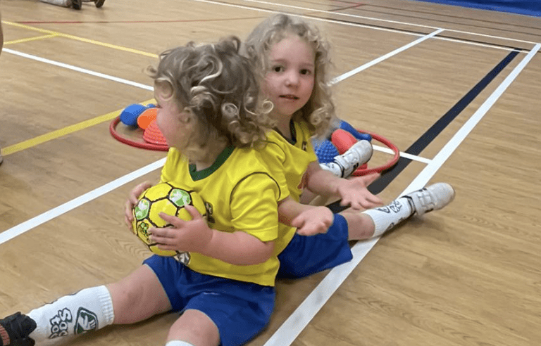 Social benefits of football. Images shows two children sitting back to back holding a baby football in a Socatots class.