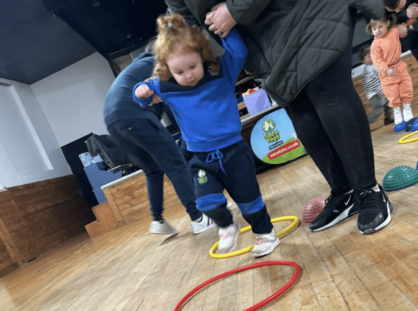 Toddlers physical development. Image shows a toddler holding hands with their parent as they walk over the coloured hoops in their Socatots football class.