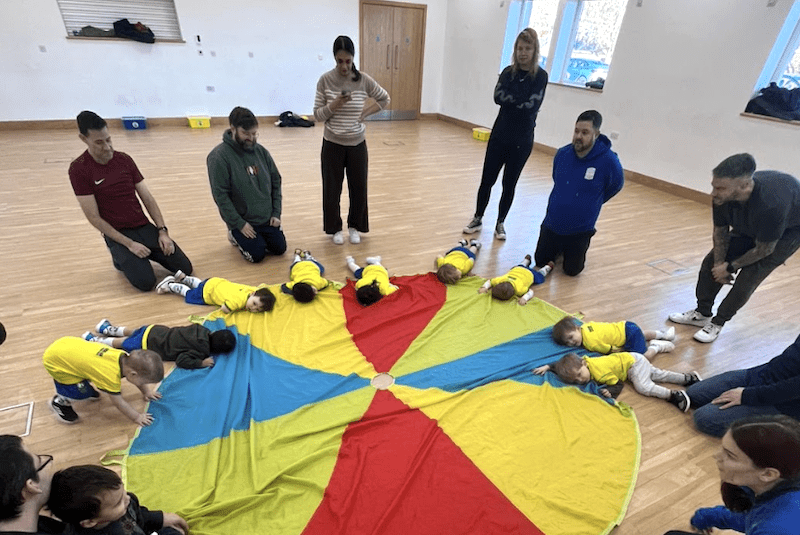 Toddler football club Dunstable. Image shows toddlers laying down on the multi coloured parachute during a Socatots class, with parents behind.