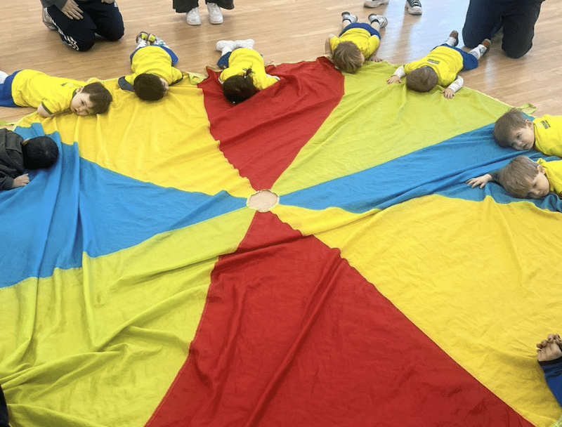 Toddler football club in York. Image shows a group of toddlers during a Socatots class laying down on the multicoloured parachute.
