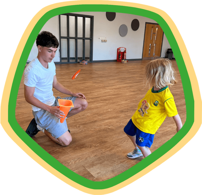 Image shows a dad and his son during a Socatots class. The dad is holding a plastic cone while his toddler kicks a beanbag into it