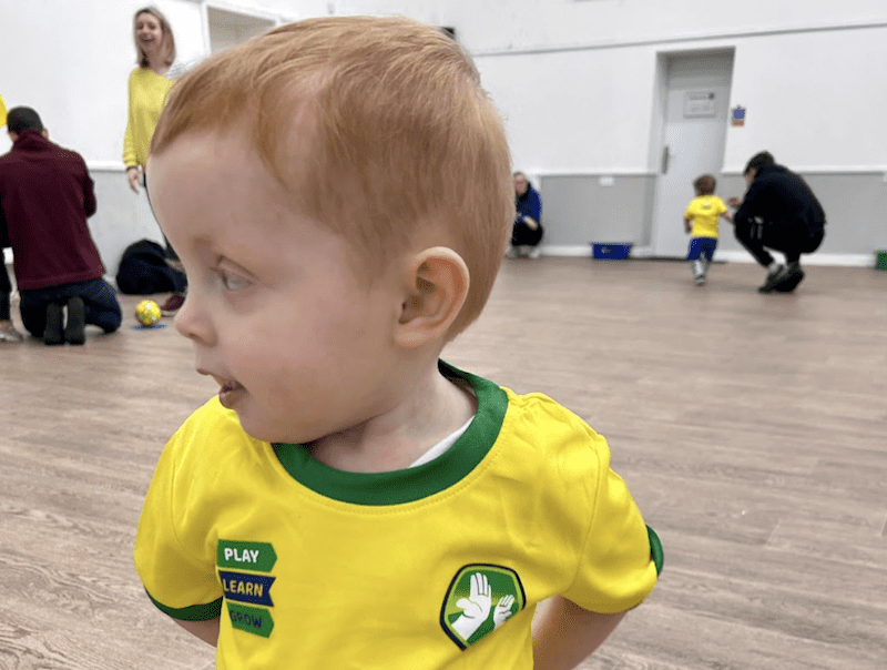 Toddler football classes in Staffordshire. Image shows a toddler in uniform at their Socatots class with a parent and toddler in the background.